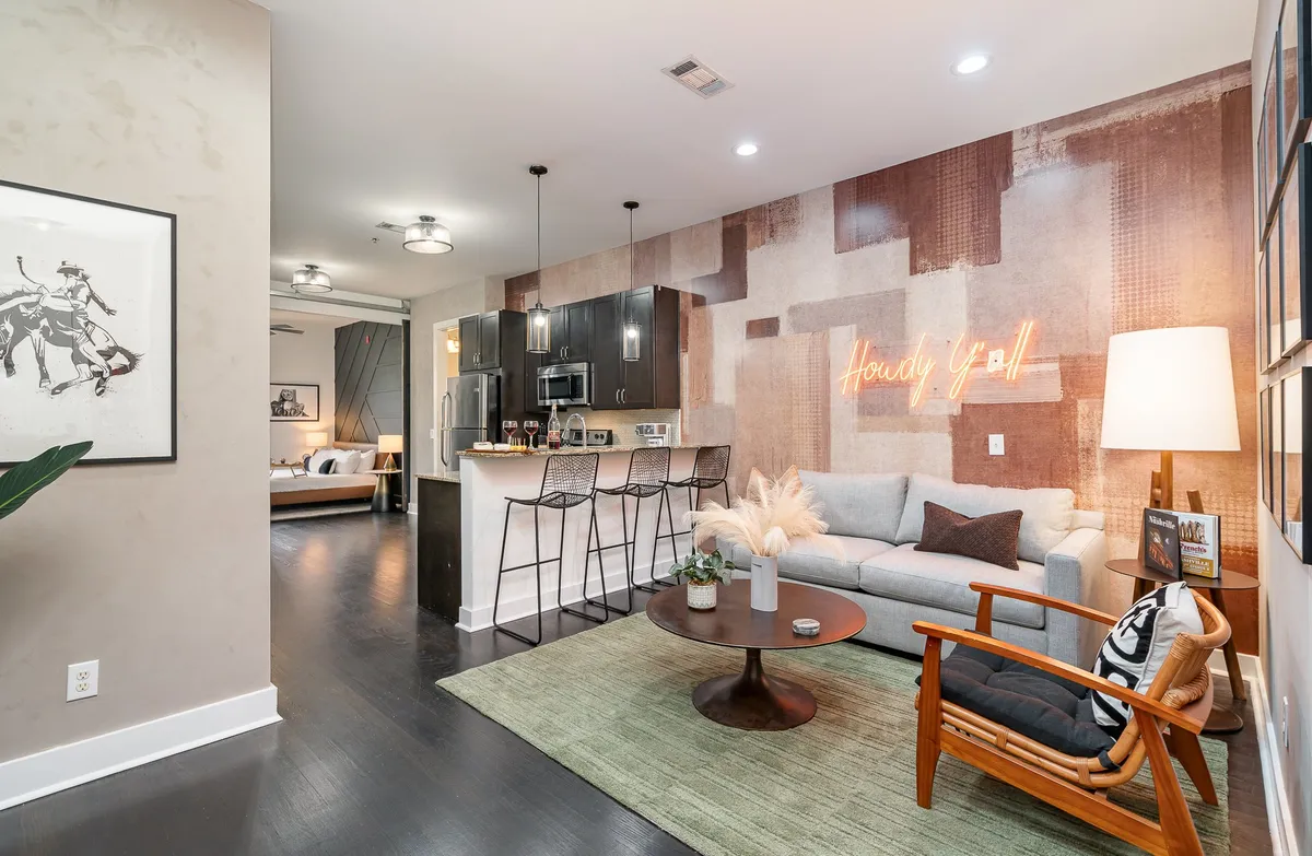 The hero image showcases a bright, open-concept living space with a vibrant 'Howdy Y'all' neon sign, a cozy seating area with plush couches, a stylish kitchen bar, and a touch of greenery on the coffee table. The scene exudes warmth and a playful Nashville vibe, perfect for gatherings or relaxing after exploring the city.