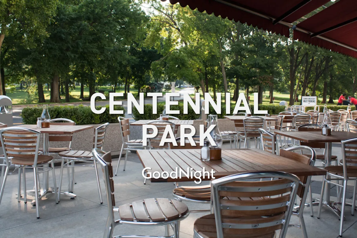 An inviting outdoor dining area featuring wooden tables and chairs set under a canopy awning, surrounded by lush greenery. The scene is casual, with space for groups to gather and enjoy meals or drinks in a lively, open-air environment.