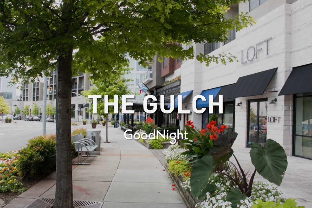 A vibrant exterior streetscape with a tree-lined sidewalk, colorful flower beds, storefronts, and benches. The scene captures people strolling, relaxing, and socializing, evoking a sense of neighborhood charm and local exploration.
