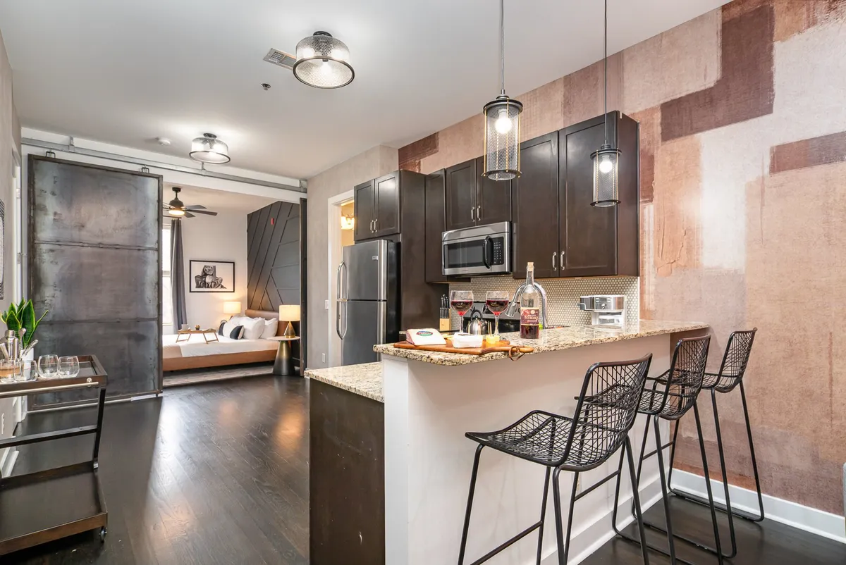 A contemporary open living space featuring a kitchen island with sleek bar stools, artistic wallpaper, a sliding barn door, and a seamless transition to the living area. The space is bathed in natural light, creating an inviting area perfect for entertaining or relaxing.