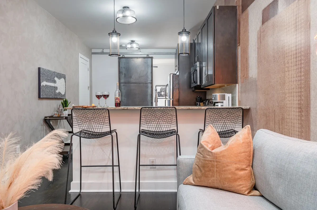 A sleek, modern interior featuring a kitchen island with bar stools, a comfortable sofa, and stylish lighting. The space is designed for casual dining, entertaining, or relaxing with friends after a night out.