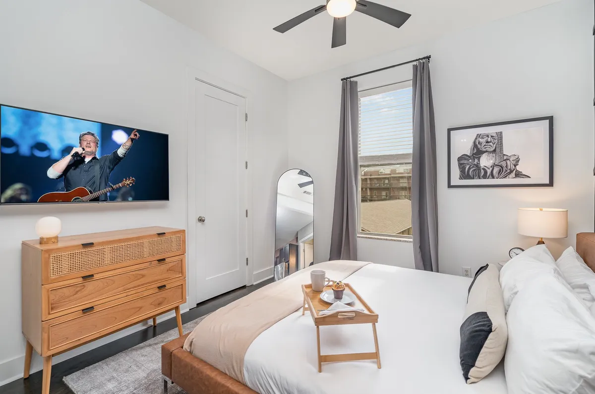 The image shows a cozy, modern bedroom featuring a comfortable bed, a TV displaying a concert scene, a nightstand, a large mirror, and artwork. The setup exudes comfort and is ideal for unwinding after a day of adventure.