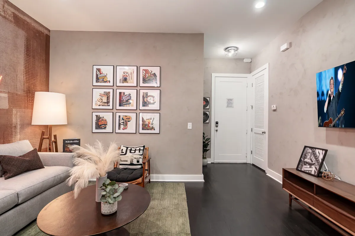 A contemporary living room scene with a sofa adorned with decorative pillows, a round coffee table, stylish framed artwork, a television (displaying an image), and a lush floor plant. The mood is both comfortable and stylish, suitable for relaxing or watching TV together.