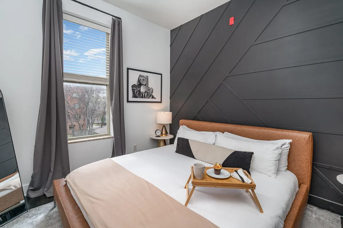 This image features a bed with a stylish headboard, a tray with breakfast, tasteful artwork on the wall, and a window streaming natural light. The ambiance is inviting and comfortable, designed for moments of relaxation, reading, or enjoying breakfast in bed.
