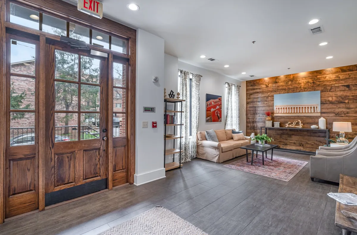 A bright and stylish building lobby featuring a large wooden front door, comfortable sofa, elegant artwork, and a welcoming plant centerpiece. The space exudes a sense of hospitality and professionalism, making a strong first impression for residents and guests.